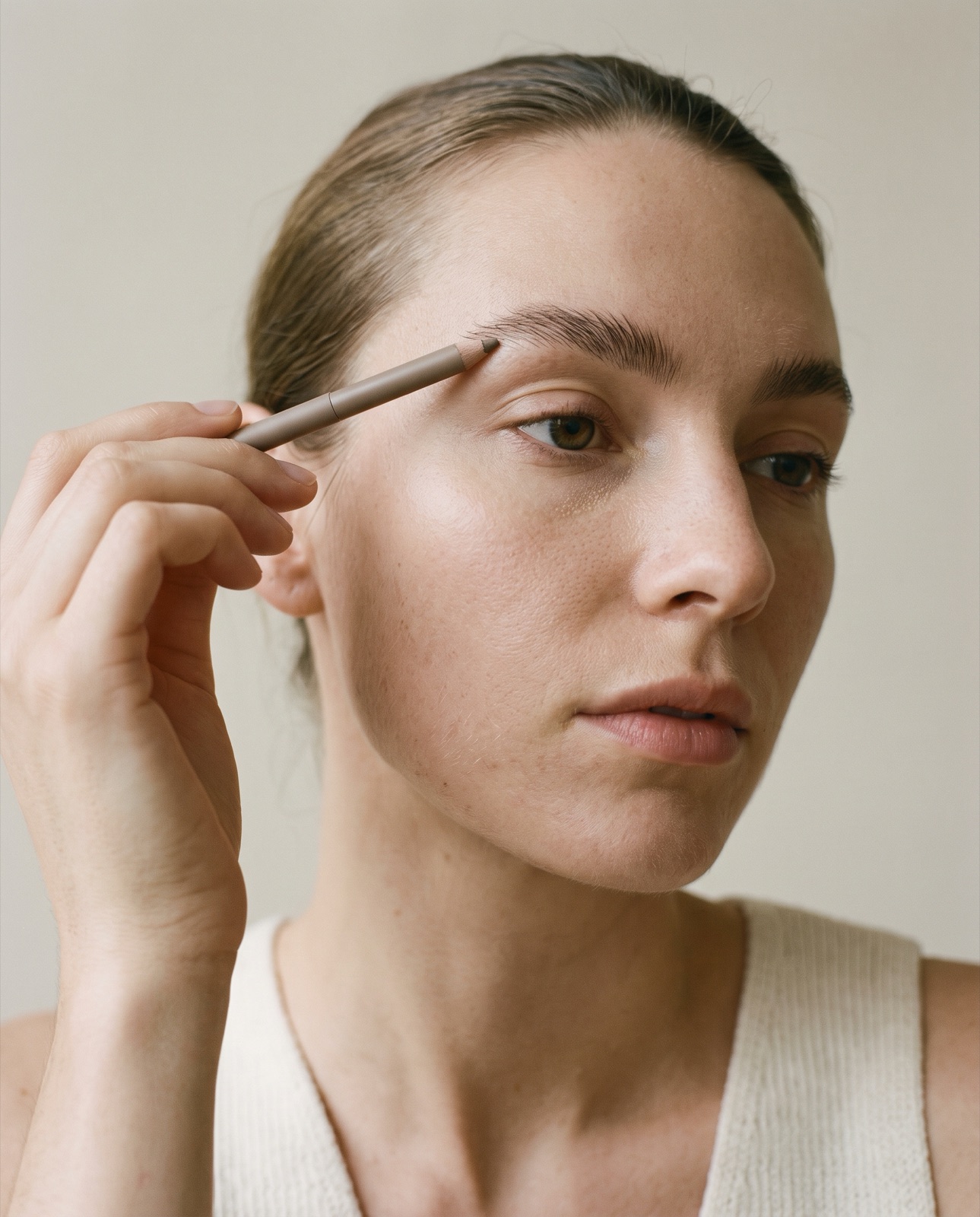 Primer plano de cejas naturales peinadas hacia arriba y una mano con un lápiz de cejas fino
