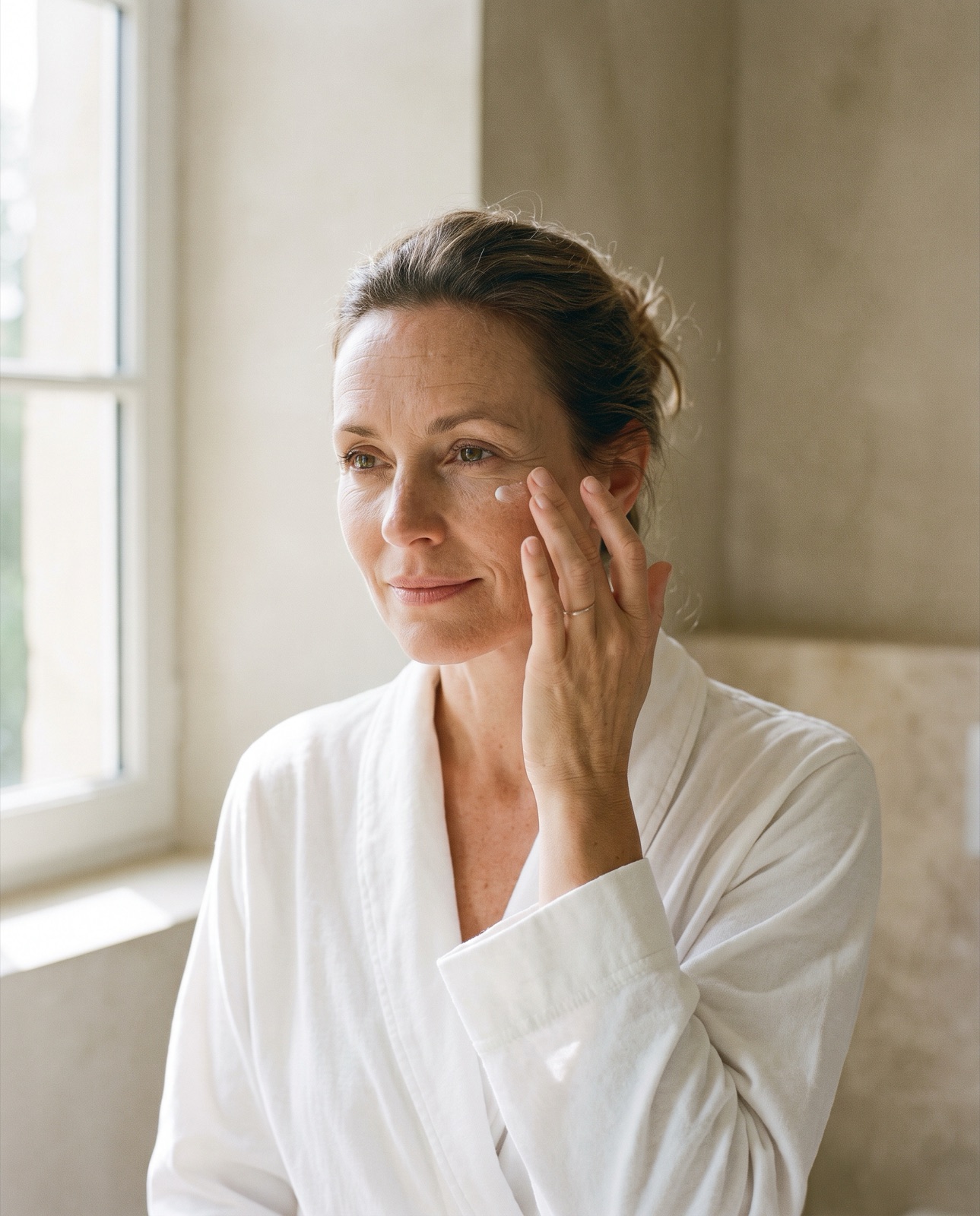 Frau tupft mit dem Ringfinger Augencreme unter das Auge, weiches Morgenlicht