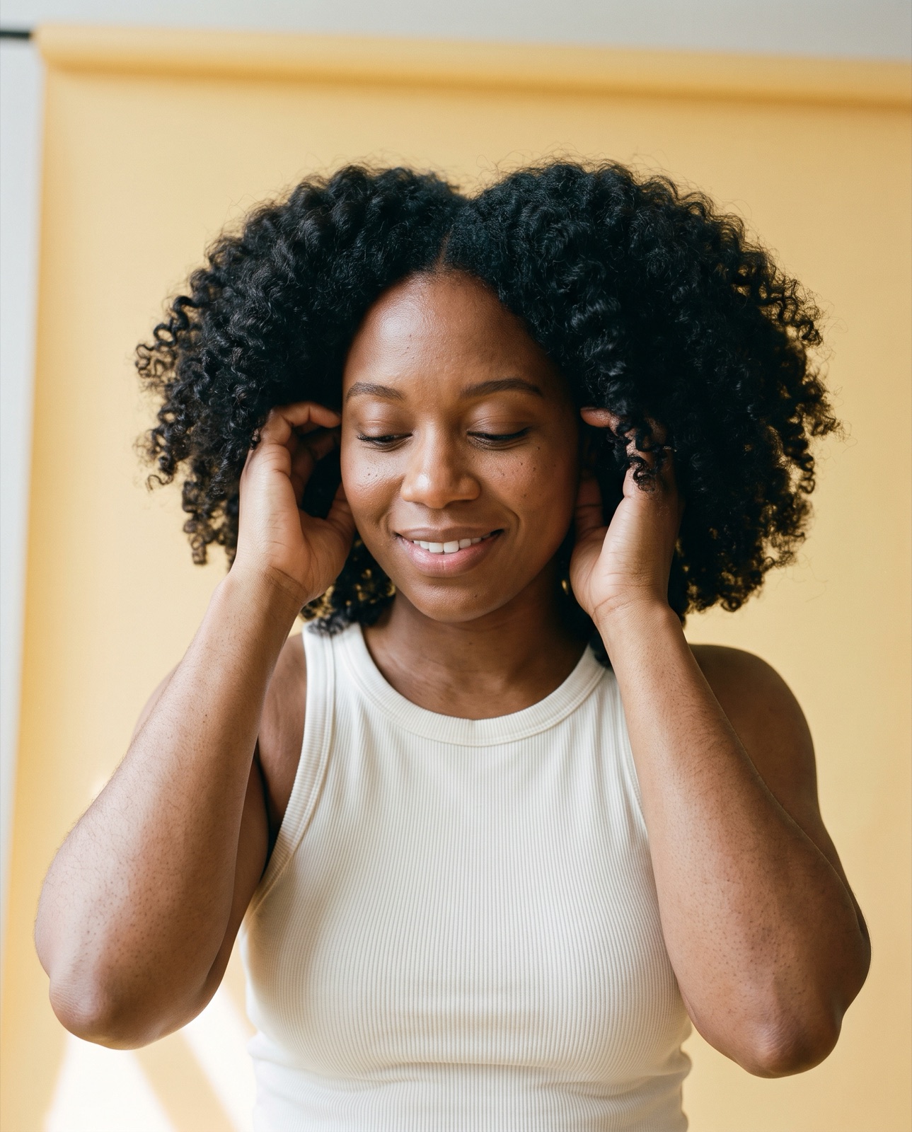 Mulher de caracóis volumosos e definidos amachuca o cabelo com as duas mãos, fundo amarelo