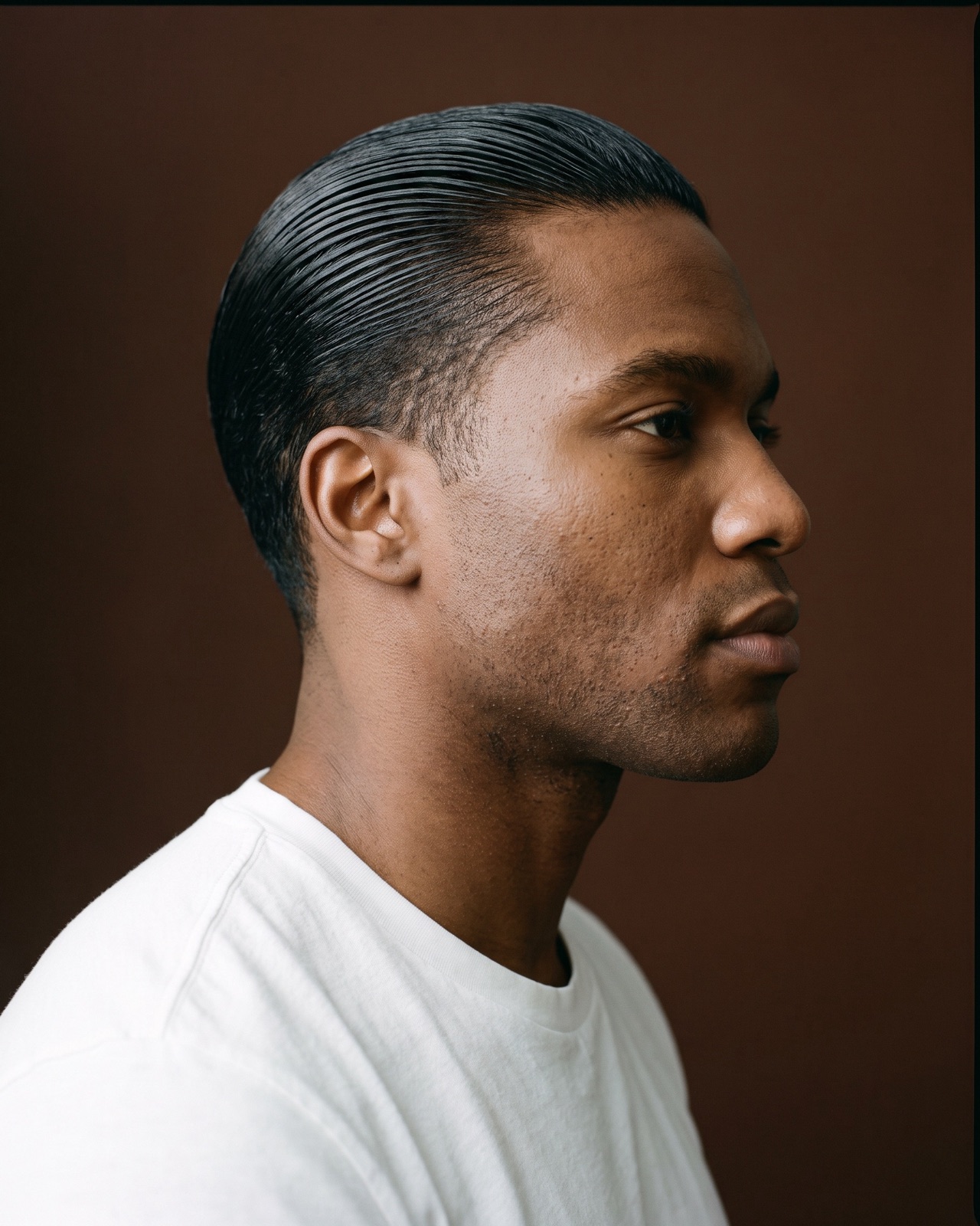 Hombre de perfil con el pelo corto peinado hacia atrás, brillante y con efecto mojado, fondo marrón oscuro