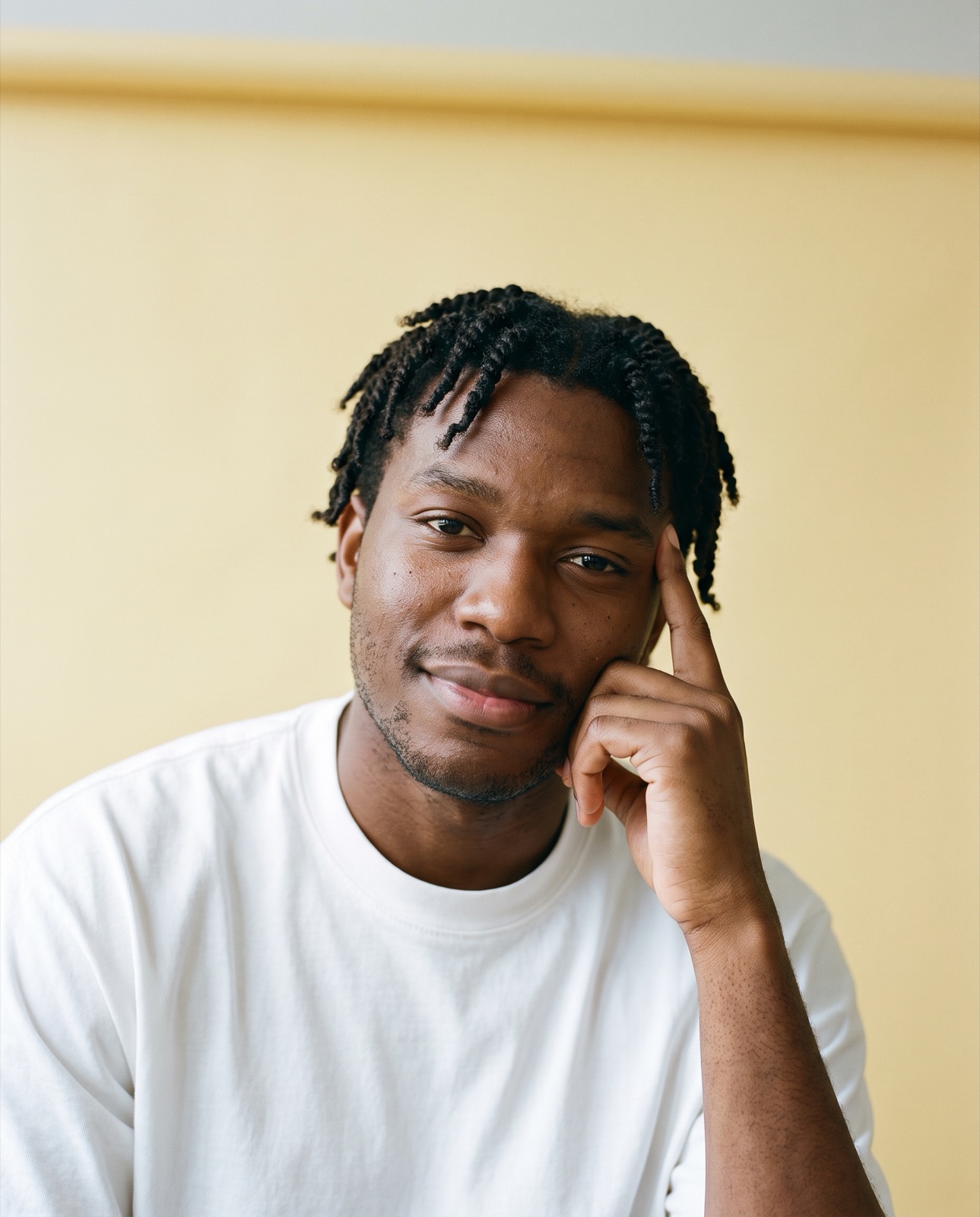 Hombre de unos treinta años con twists cortos, leve sonrisa, yemas en la sien, fondo amarillo