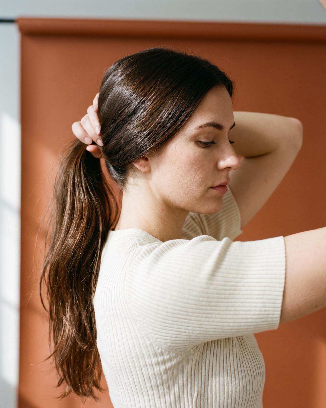 Woman in profile gathering her long glossy dark brown hair into a low ponytail with both hands